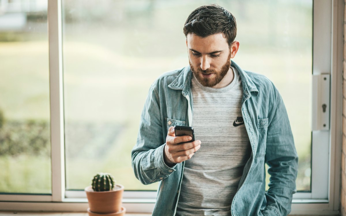 Homem consultando o celular em pé junto a uma janela